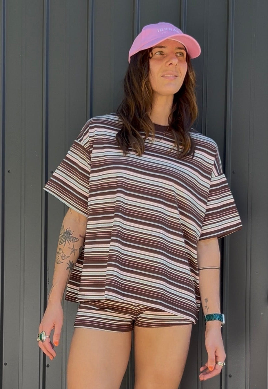 Person wearing a striped shirt and shorts with a pink cap against a gray wall.
