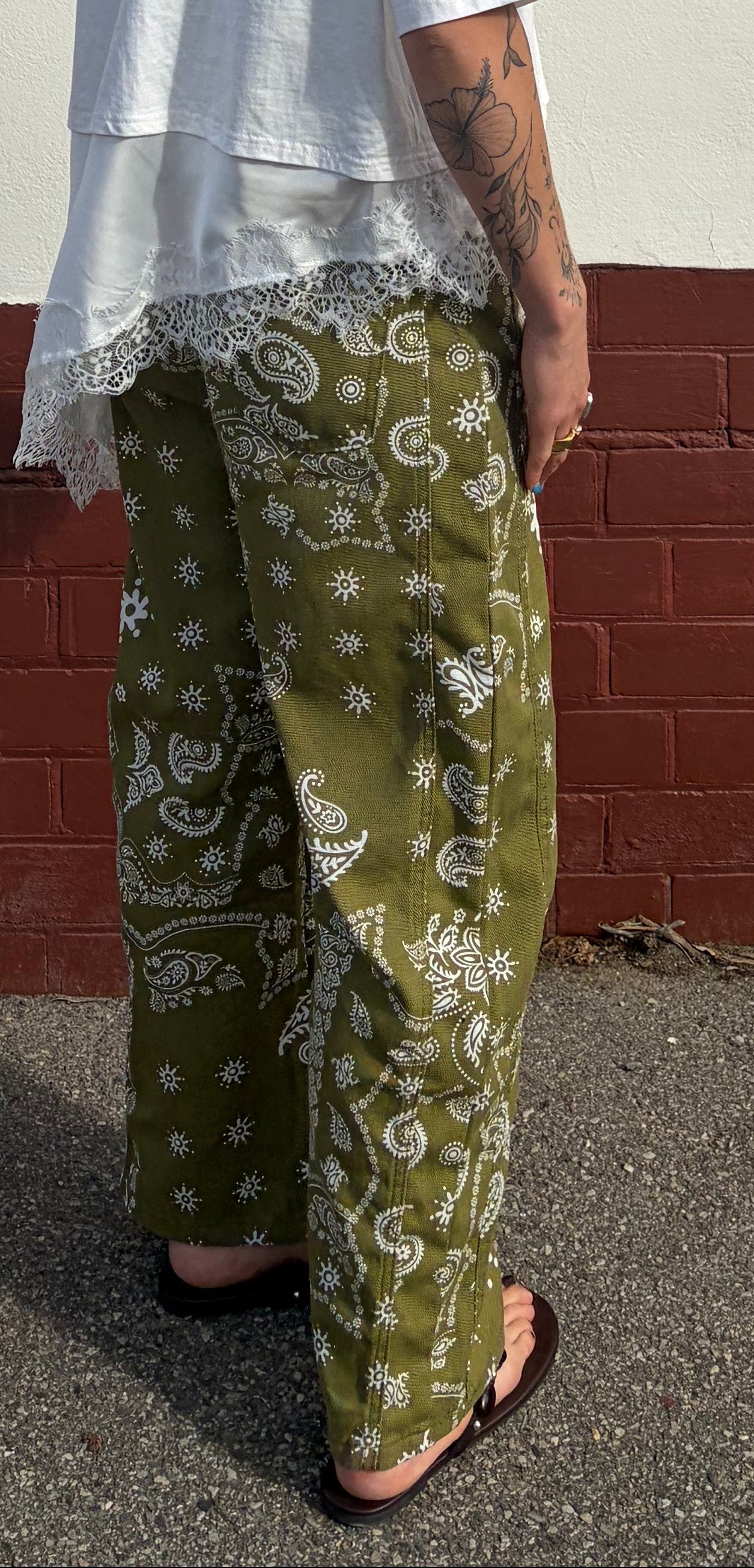 Person wearing green patterned pants and a white top against a red brick wall.