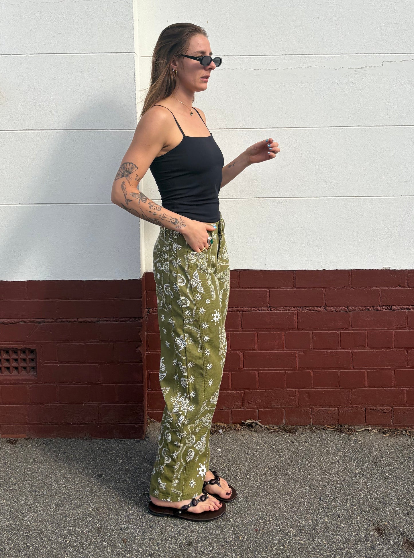 Woman wearing a black tank top and green floral pants standing against a white wall with a red base.